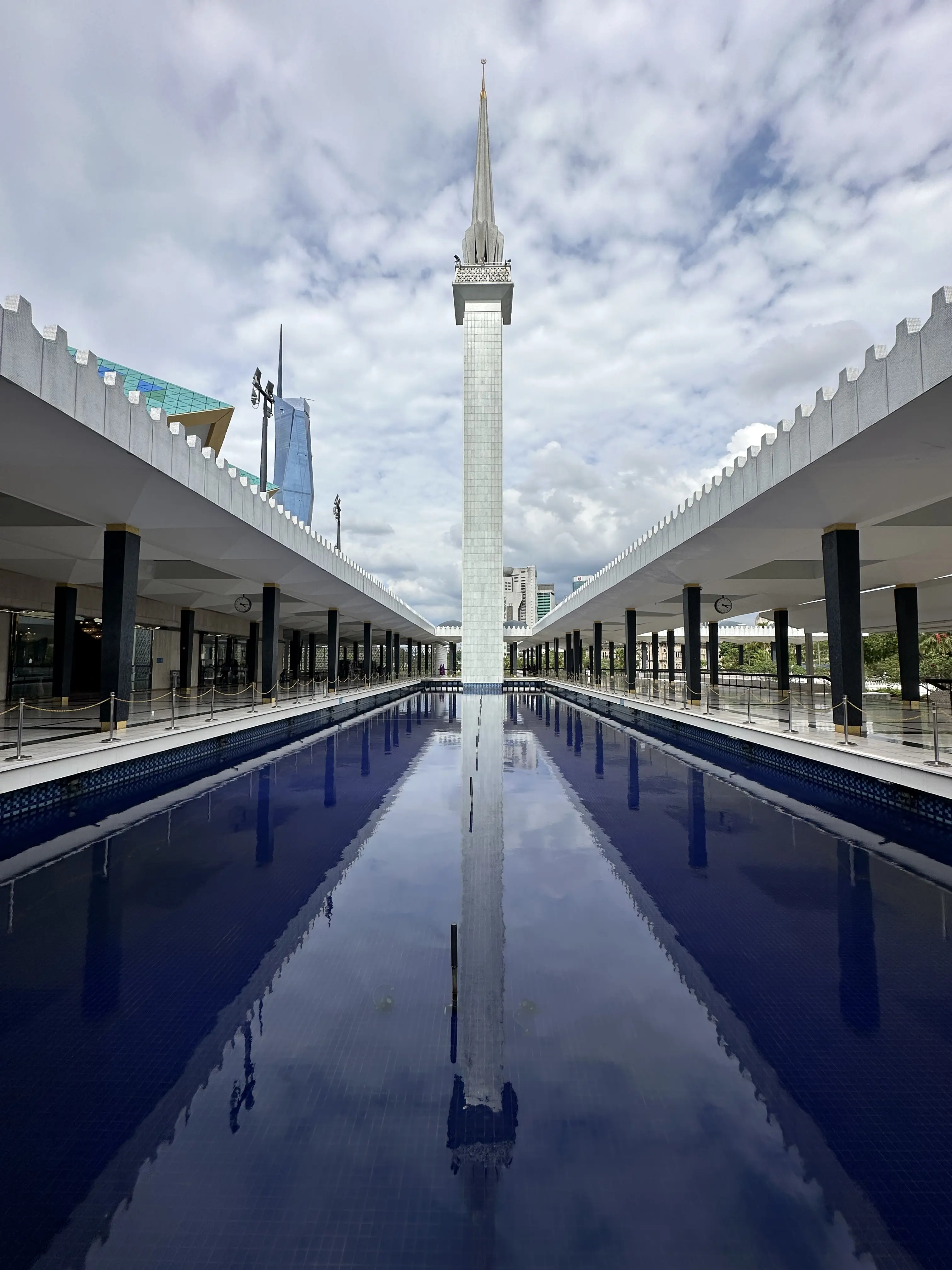 Immagine in prospettiva del minareto della Moschea Nazionale della Malesia a Kuala Lumpur. Lungo la via per il minareto, c'è una piscina colma di acqua di un azzurro brillante. Ai lati della piscina, due corridoi con numerose colonne nere e un tetto bianco