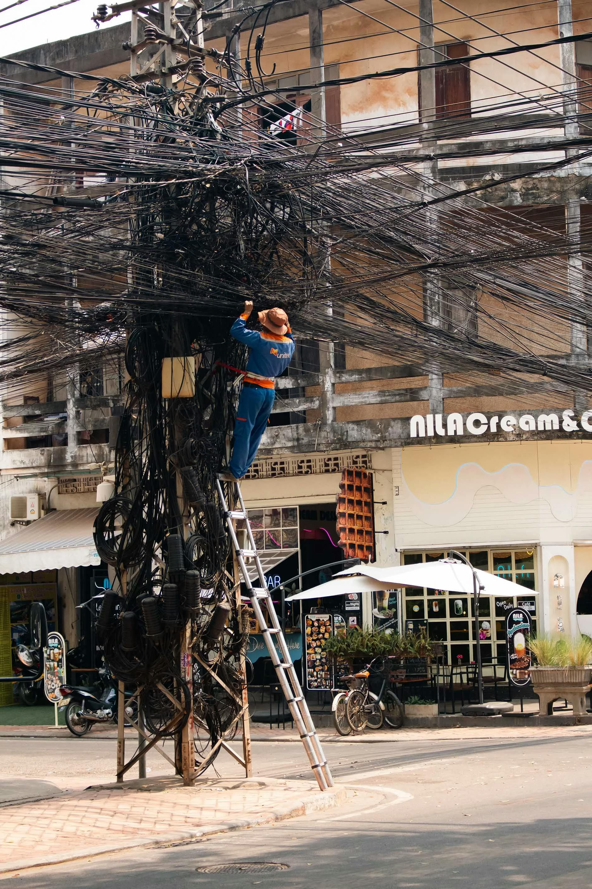 a man on a step ladder working with a lot of cable in Vientiane (Laos). The man is wearing a yellow helmet and a blue shirt