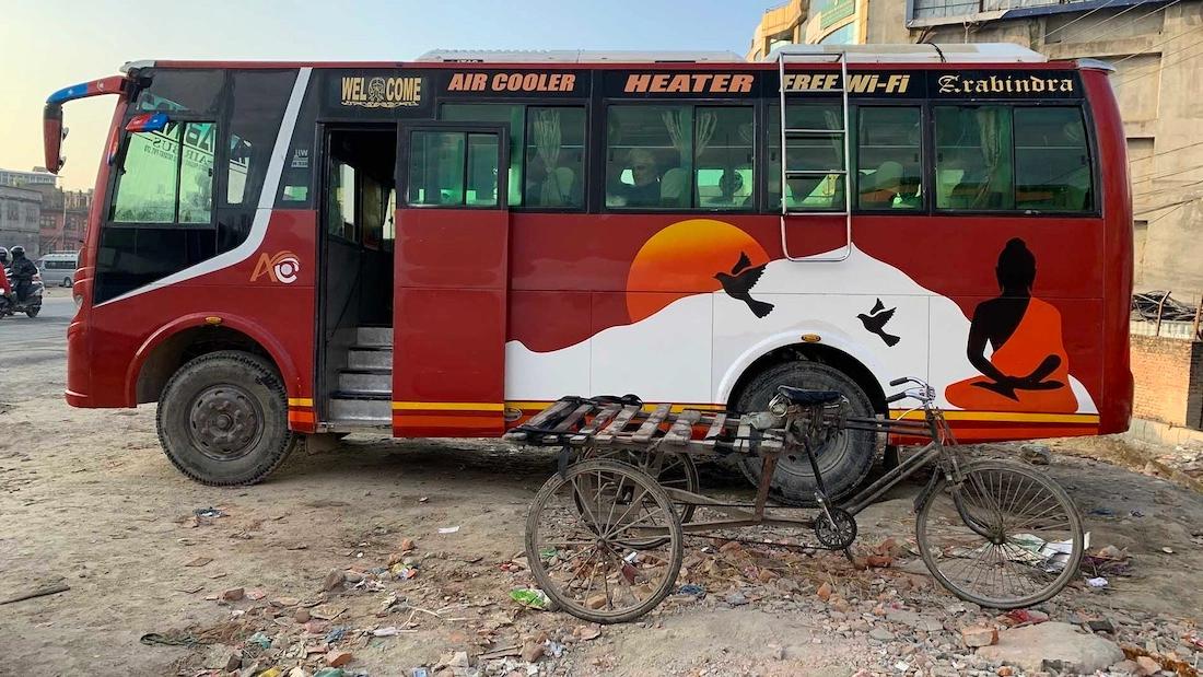 Old red bus on side of the road with a tricycle in front. The ground is dusty with rubbish and leaves. The bus has signs like "air cooler", "free wifi", "heater", "welcome"