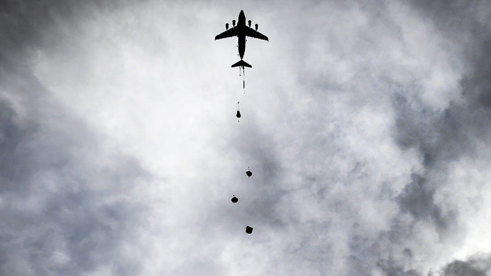 In alto, orizzontalmente al centro, si trova un aereo da caccia che sta sganciando bombe. Il cielo è coperto da nuvole scure. La foto è in bianco e nero.