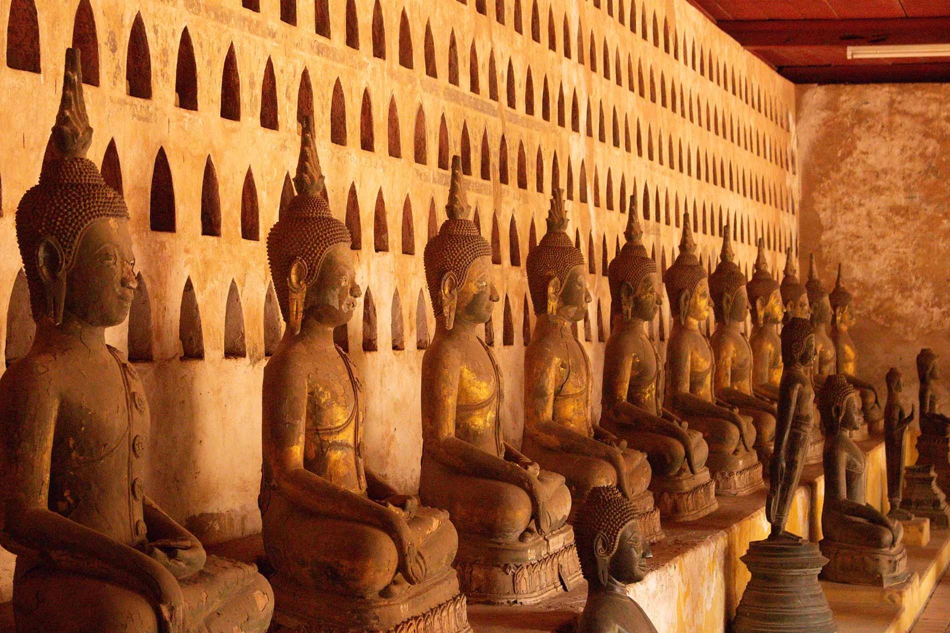 Immagine in prospettiva di alcune statue di Buddha in un tempio a Vientiane, Laos. Le statue rappresentano Buddha in una posizione seduta e sono tutte di un colore che varia dall'arancione al marrone. La parete dietro le statue ha molti "buchi" contenenti statue di Buddha più piccole, che non sono visibili in questa immagine. Su un livello inferiore ci sono alcune statue di Buddha più piccole, in bronzo, in pose diverse.