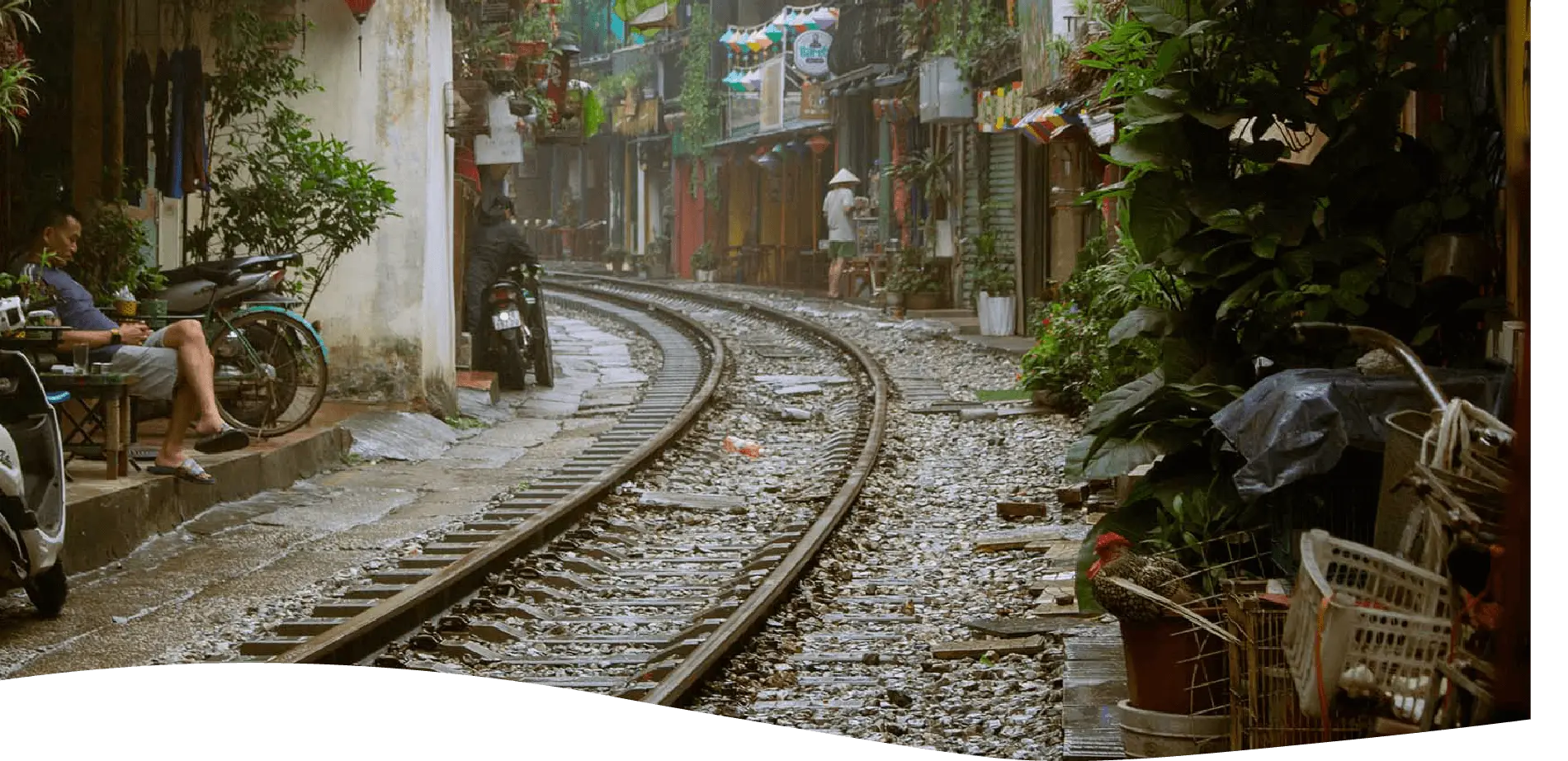 hanoi train street, on the left there is a man enjoying his coffee, while on the background we can see a a lot of plants hanging from the baclony of houses, which are right nect to the railtrack