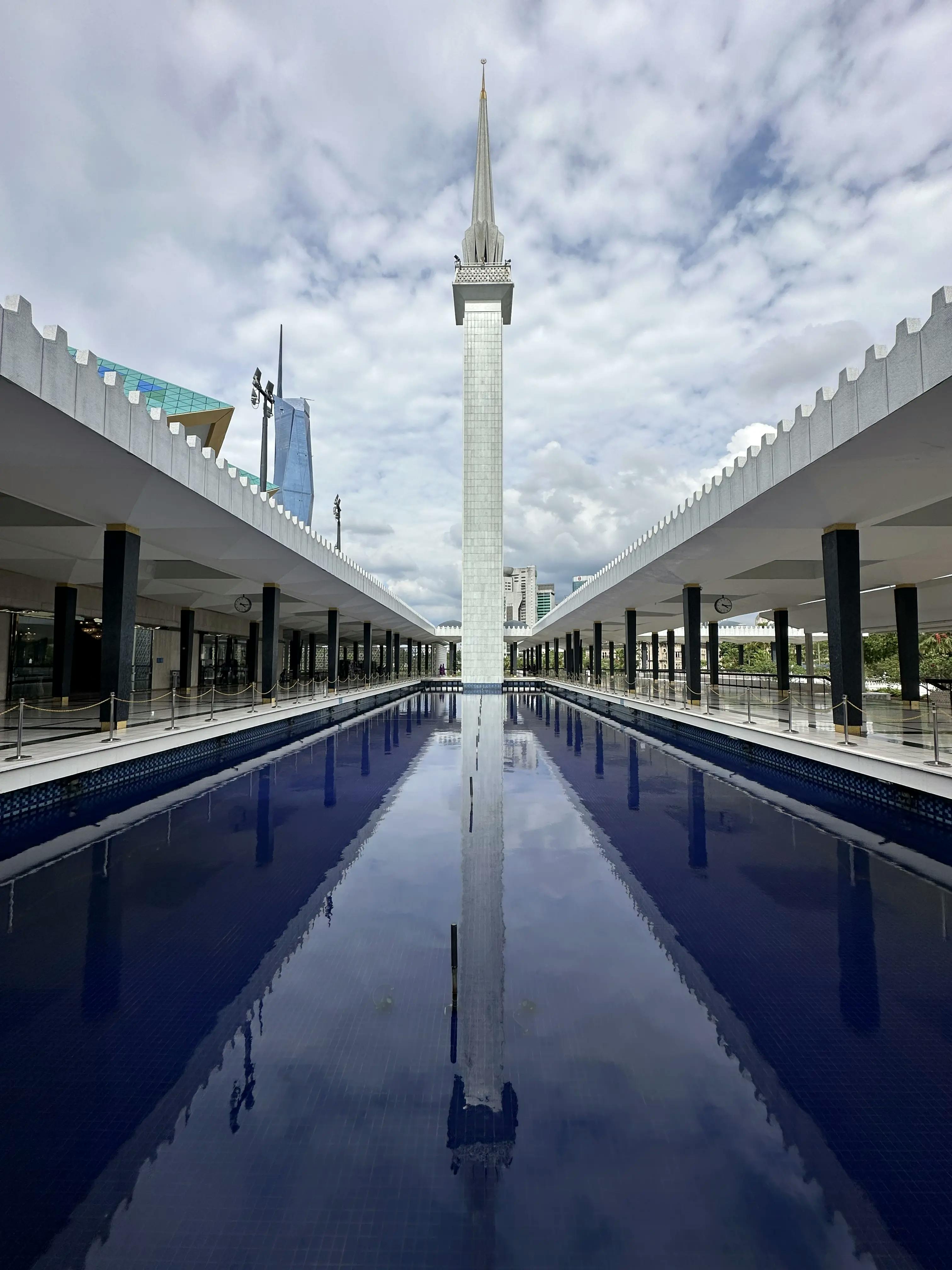 picture in perspective of the minaret of the National Mosque of Malaysia in Kuala Lumpur. On thw way to the minaret there is pool full of bright blue water. On the side of the pool, two corridors with a lot of black columns and a white roof