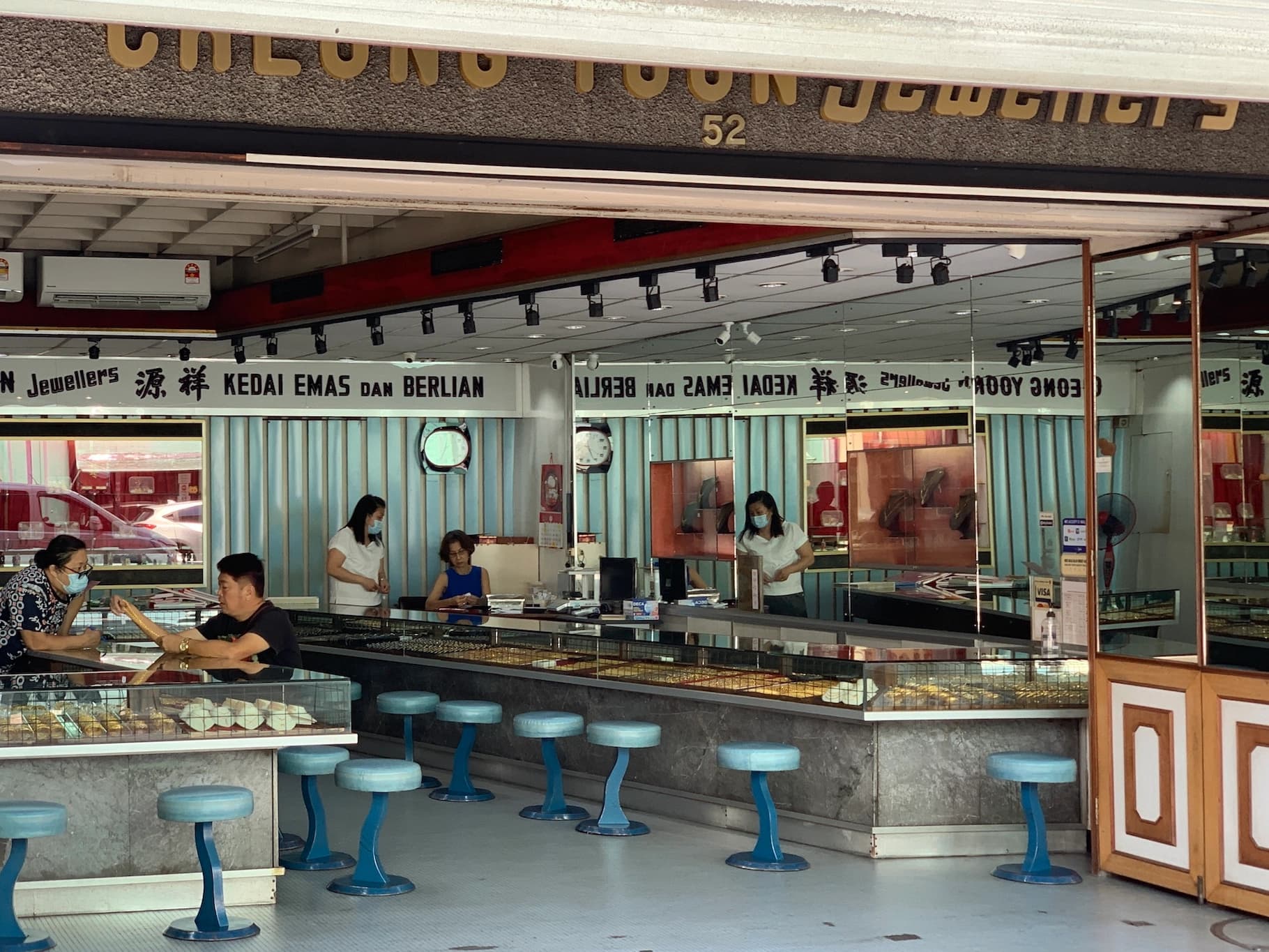 missing image Scena di vita quotidiana in un negozio d'oro in Malesia. Il negozio presenta due banconi opposti, ciascuno con numerosi oggetti d'oro esposti. Sotto il bancone sulla destra ci sono alcune piccole sedie rotonde, colorate di azzurro chiaro. All'interno del negozio ci sono alcune persone. A destra ci sono due commesse che parlano tra loro, mentre a sinistra c'è un cliente con una collana d'oro in mano, che sta parlando con un altra commessa.