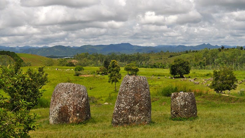 Three jars in a line from the Plain of jars. The jars sit on green grass where you can see some trees scattered around. In the background, some hills and the sky, covered by clouds 