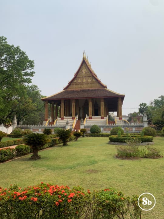 Former temple of Haw Phra Kaew, Vientane The photo gives a glimpse of Haw Phra Kaew, a former temple located in Vientiane, Laos. In front of the temple, there's a garden with grass, and here and there, there are also some small trees. In the foreground, there's a hedge with red flowers. The temple is in the background. The temple is viewed from the front, with the upper part colored in gold, and the roof colored in red. Below the roof, there are many golden columns.