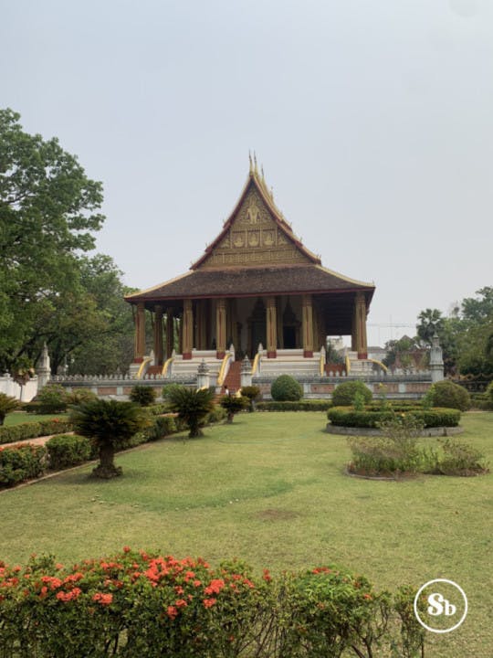 The photo gives a glimpse of Haw Phra Kaew, a former temple located in Vientiane, Laos. In front of the temple, there's a garden with grass, and here and there, there are also some small trees. In the foreground, there's a hedge with red flowers. The temple is in the background. The temple is viewed from the front, with the upper part colored in gold, and the roof colored in red. Below the roof, there are many golden columns.