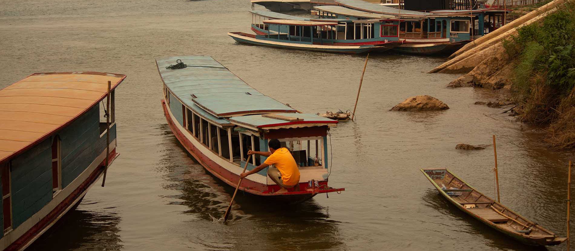 Un uomo nel retro della sua barca a lunga coda, con un remo in mano, che sta spingendo la propria barca sul fiume Mekong. Foto scattata nei pressi di Luang Prabang, nel Laos