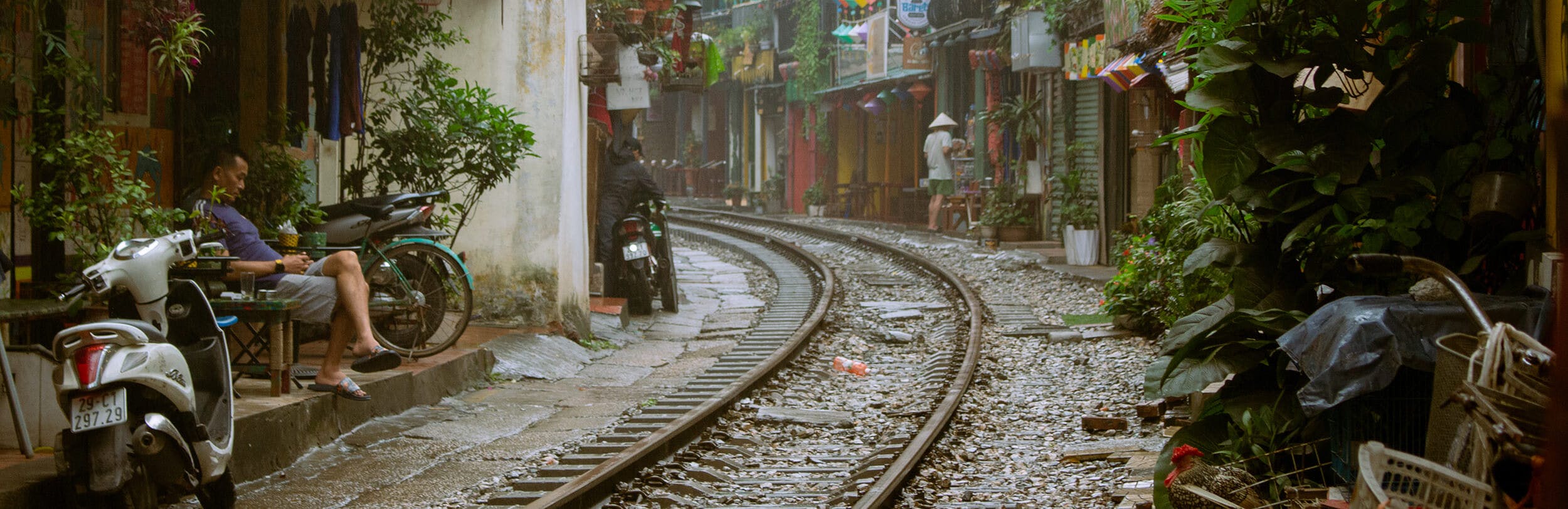 Picture of Train Street, Hanoi (Vietnam). There is a slight drizzle in the air. On the left a man is drinking some coffee, or tea; a little bit further up, another person is trying to cover their scooter from the incoming rain. There is a lot of green plants all around 