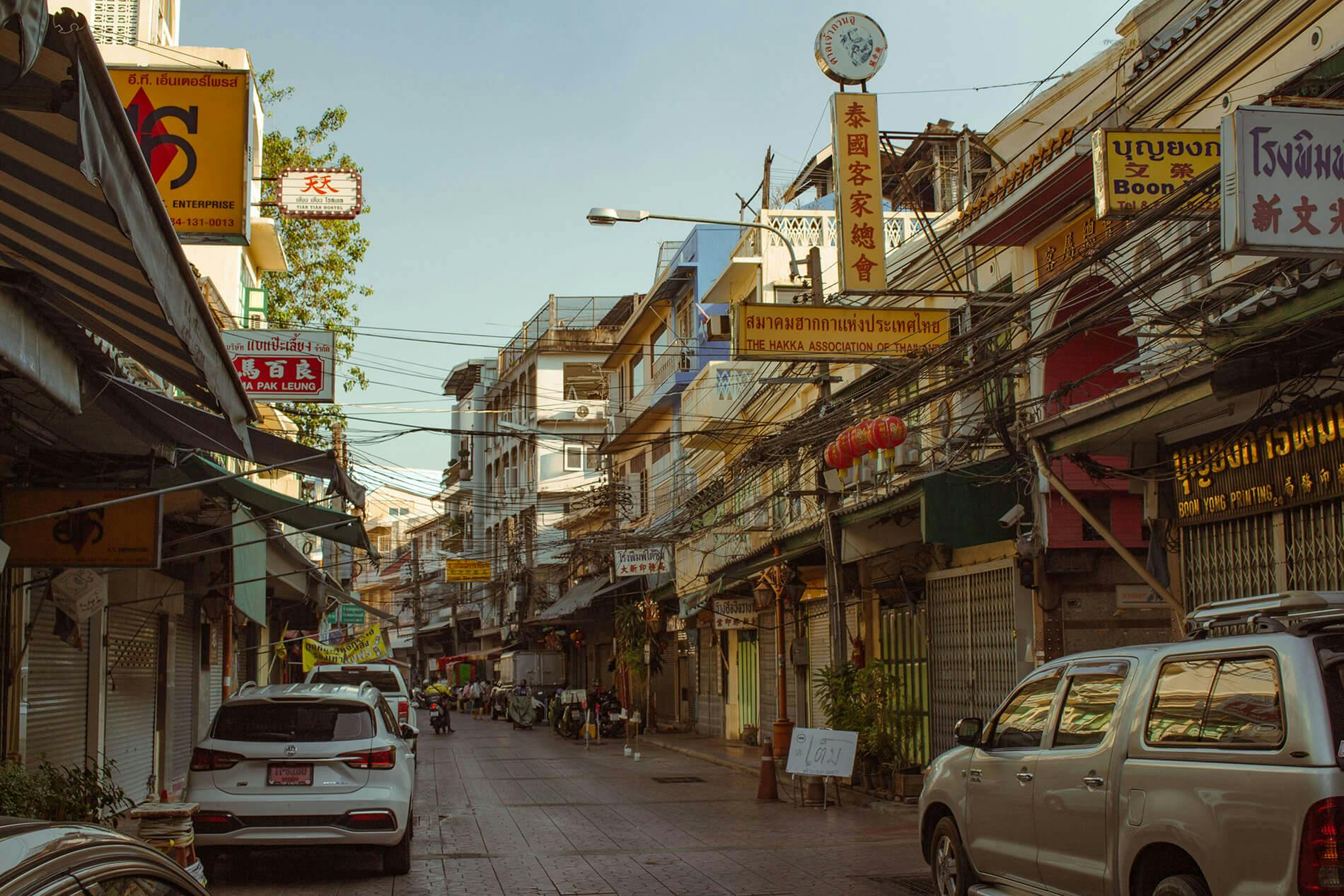 Image of Chinatown, Bangkok. There are a few cars on the road and many signs from various shops. Most signs are in Chinese and they are yellow with the text in a bold red