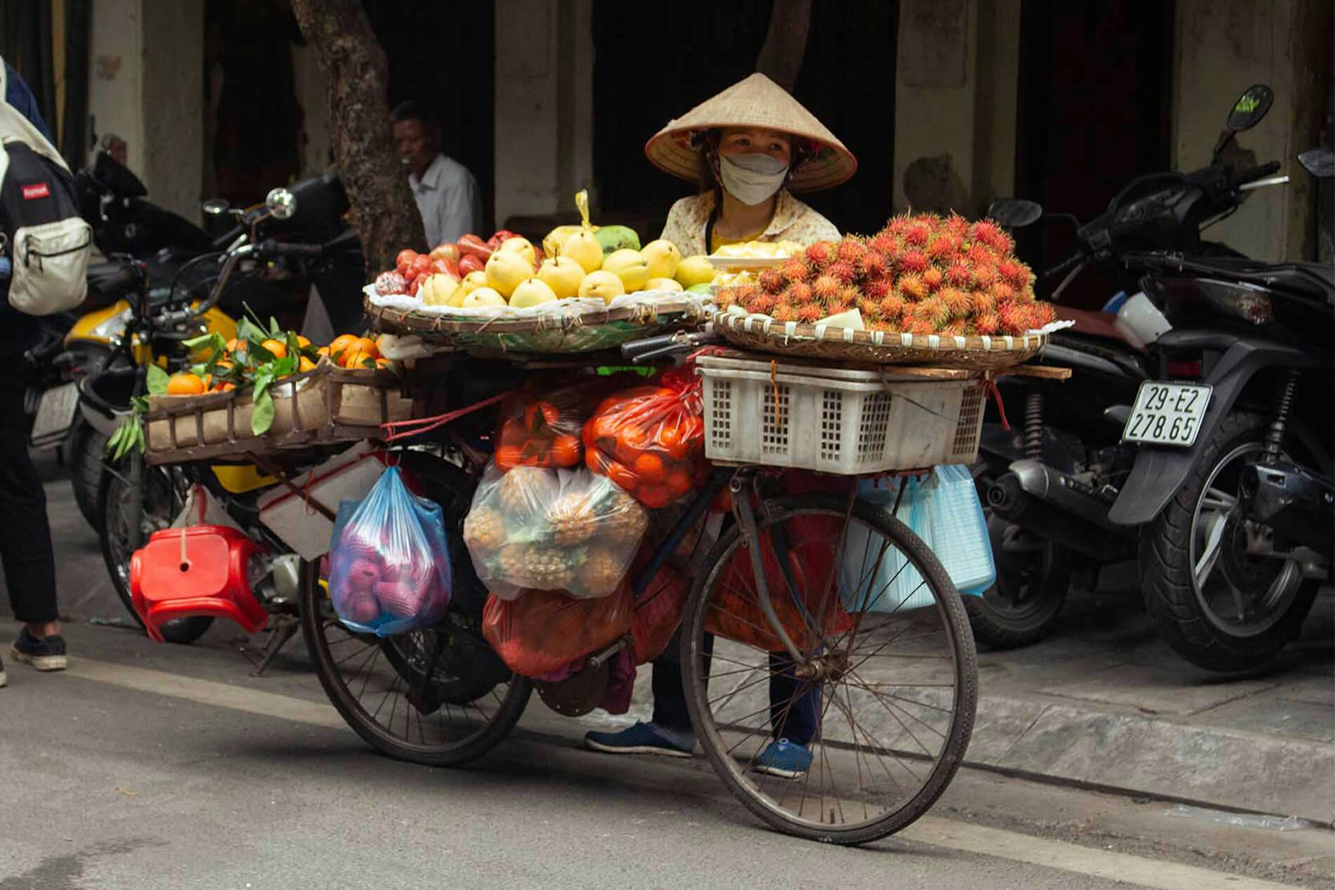 A woman in Hanoi is carrying a lot of different fruits with her bike. She is standing behind the bike. The woman is wearing a traditional Vietnamese conical hat, and she's also wearing a mask. The fruit is very colourful, with yellow, orange and red being the most visible colours.