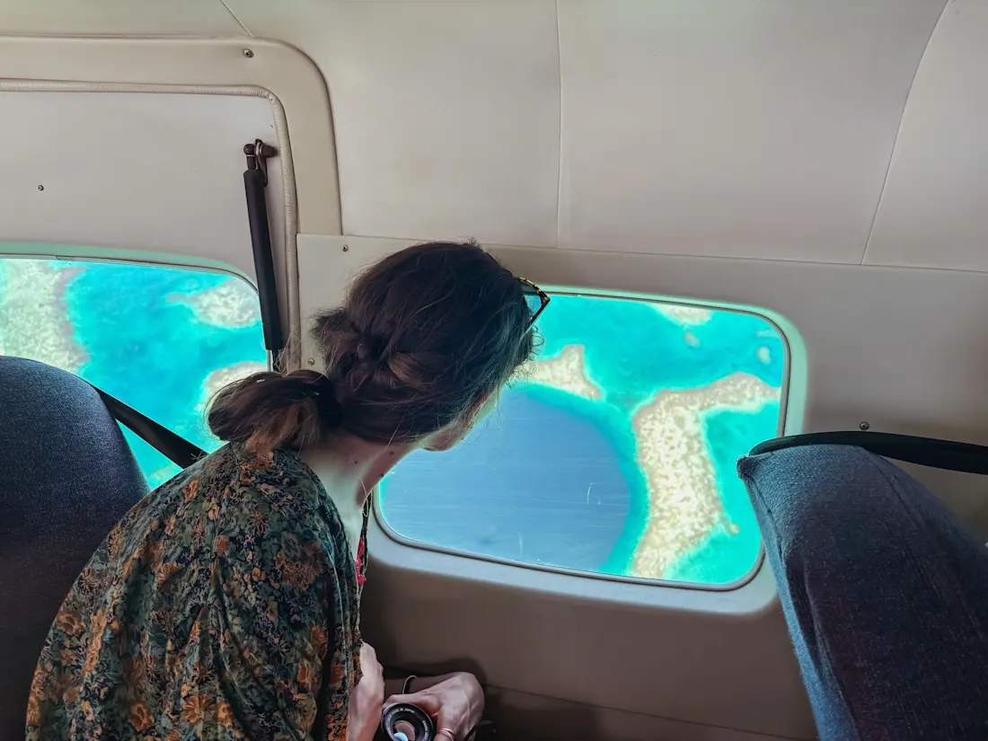 The Great Blue Hole Belize Central America Voragine in blu intenso, perfettamente circolare, su una barriera corallina vista dal finestrino di un piccolo aereo. Una persona tiene una macchina fotografica in mano mentre guarda fuori dal finestrino.