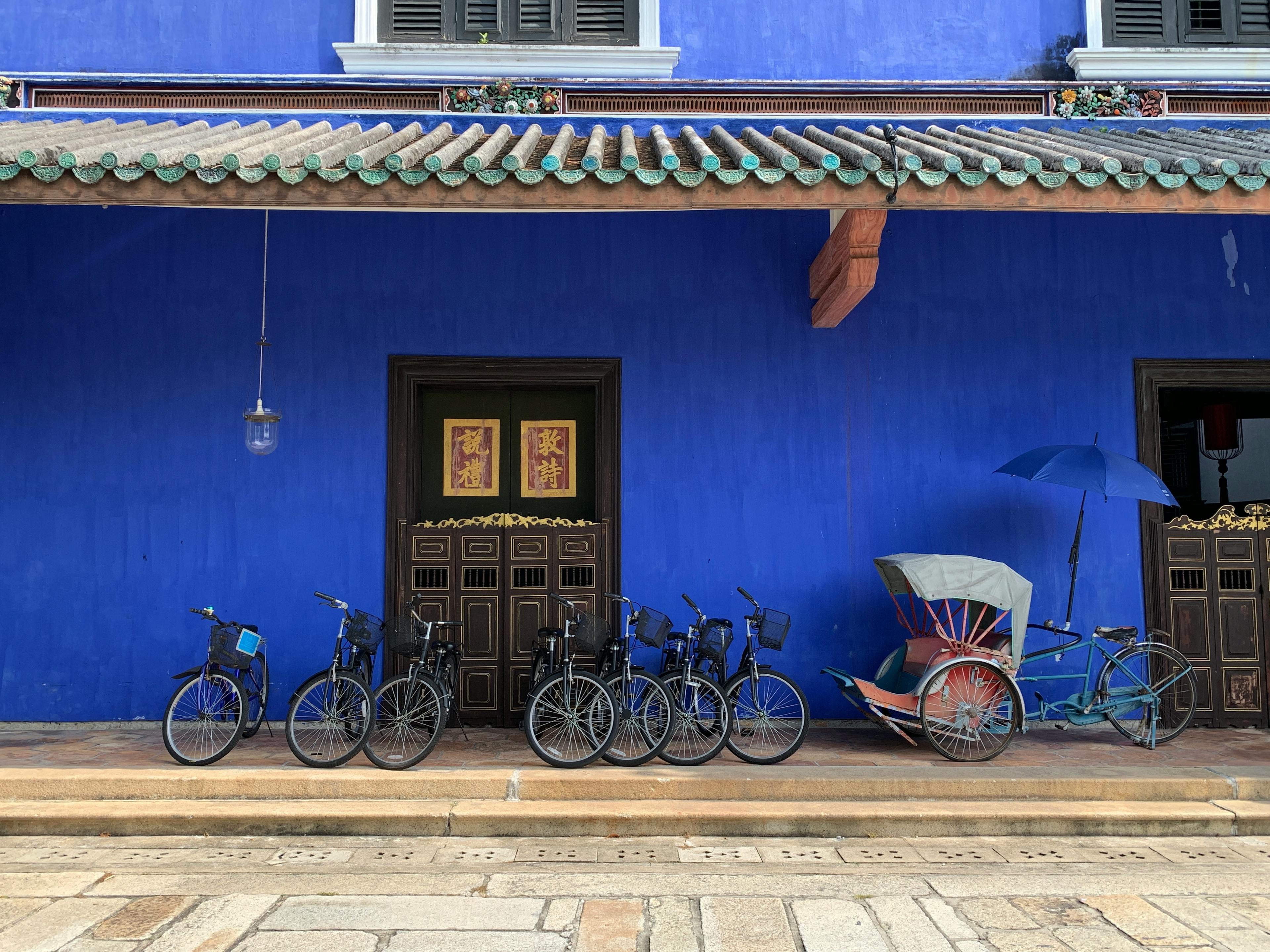 Detail of the Blue House in Georgetown, Penang. The wall is painted with a strong, bright, blue ocean. There is a black door, with a grey frame. Within the door there are two Chinese character inscribed in a wooden frame of red background, with the characters, as well as the border, painted in gold. In front of the door there a few bikes disposed on a row, and on the right end there is another bike with a rickshaw attached to it.