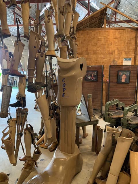 Prostheses on display at the COPE museum in Vientiane The photo has been taken at the COPE museum of Vientiane and it contains a few prosthesis. Thy are all prosthesis for legs and there is a main one in the front which stand still, posed on the floor. It also features the write "COPE" and a heart drawn in black on the right side. Around the are a few more prosthesis, all of pretty much the same color (yellowish to brown), but some of them are hanging from the ceiling.