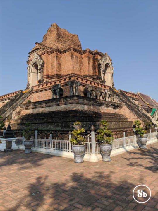 Vista di Wat Chedi Luang da uno degli angoli. Il tempio è parzialmente in rovina, ma si riescono comunque ad intravedere delle statue di elefante sia nei lati che nell'angolo. Il tempio è principalmente composto di mattoncini rossi. Nella parte superiore ci sono due archi, uno per lato, di color bianco-grigoignolo. 