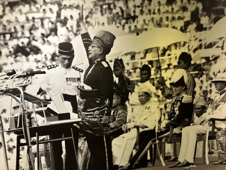 Picture, in black and white, of president Tunku Abdul Rahman proclaiming "Merdeka",  on 31 August 1957. Tunku Abdul has his right hand open and lifted towards the sky. Next to him and behind him there are a few high officials of Malaysia. In the background, a big crowd of Malaysian looking at the president.