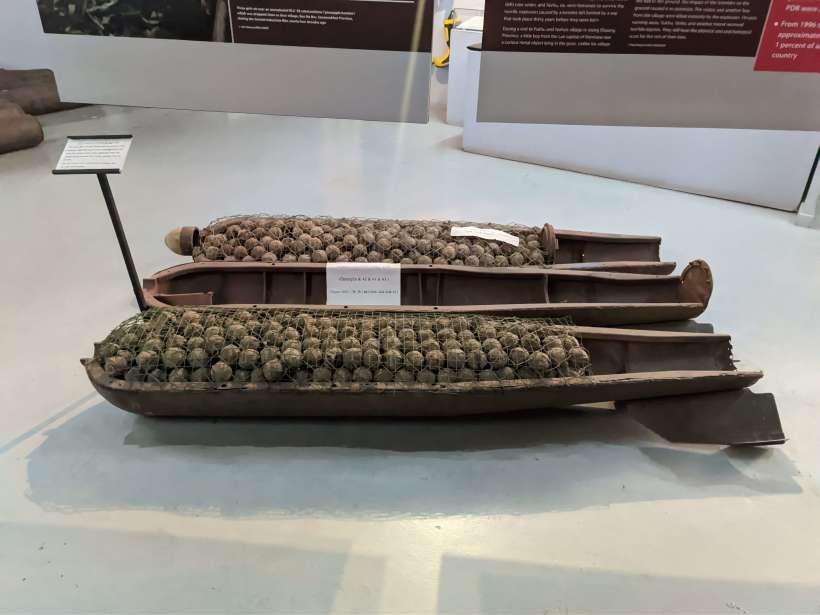 Interno di una bomba a grappolo piena di submunizioni, in mostra al museo COPE di Luang Prabang Interno di una bomba a grappolo piena di bomblets, esposta al museo COPE a Luang Prabang. La bomba è aperta e si possono vedere tre parti. Le parti sinistra e destra sono piene di bomblets, mentre la parte centrale è vuota, ma contiene un foglio con qualcosa scritto sopra. A sinistra della bomba c'è un palo con un'etichetta bianca sopra che fornisce ulteriori dettagli sulle bombe.