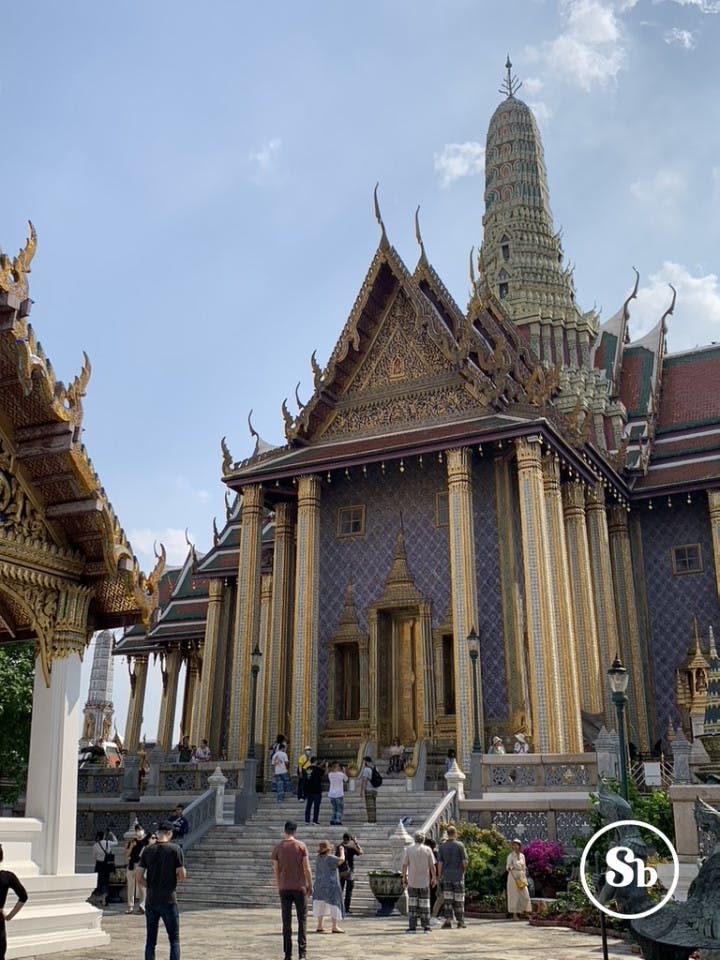 Vista del Wat Phra Kaew, dimora del Buddha di Smeraldo, a Bangkok. Nella foto si intravede la facciata del palazzo, con un muro di un acceso viola, di fronte a cui ci sono delle bellissime colonne dorate. Il tetto è di forma triangolare con un colore rosso, e dietro al tetto si intravede una stupda.
Sulle scale del palazzo ed in fronte al palazzo ci sono molti turisti che ne ammirano la bellezza, alcuni dei quali scattano anche delle foto.