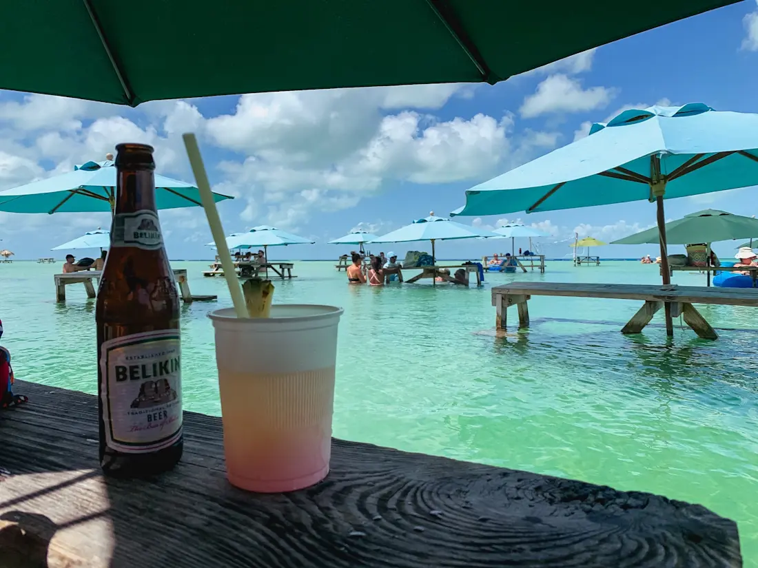 Ristorante Blue Bayou a Secret Beach Bottiglia di birra Belikin e bicchiere con rum punch su un tavolo all’ombra di un ombrellone, con tavoli da picnic mezzo sommersi nell’acqua turchese e ombrelloni aperti sullo sfondo.
